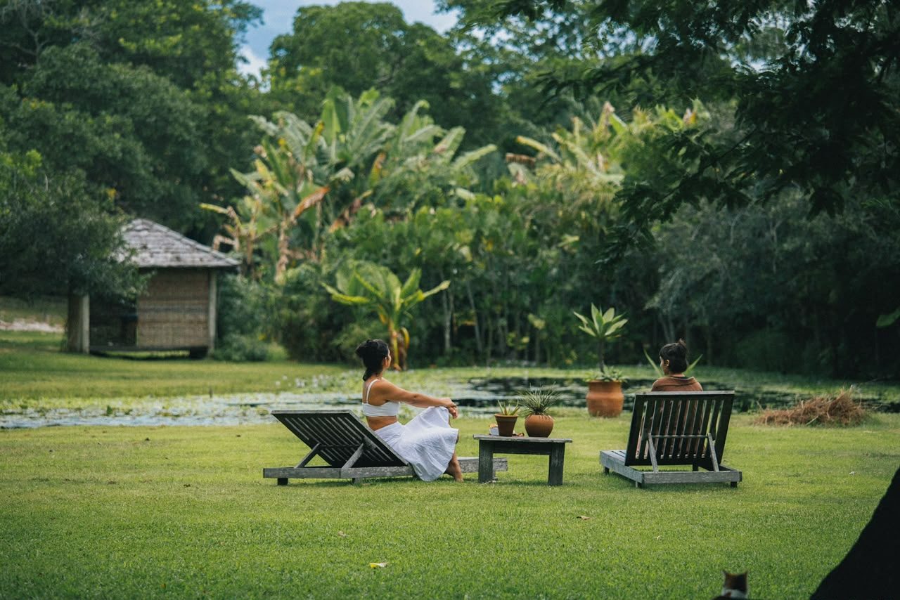 Duas mulheres relaxando em espreguiçadeiras à beira da lagoa, com bangalô e jardim tropical ao fundo
